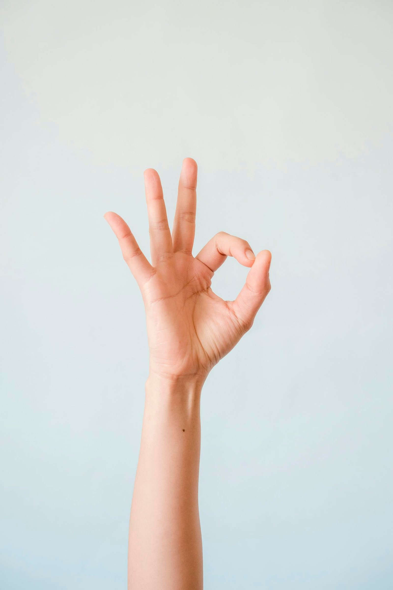 A single human hand making an OK gesture, isolated against a soft pastel background.